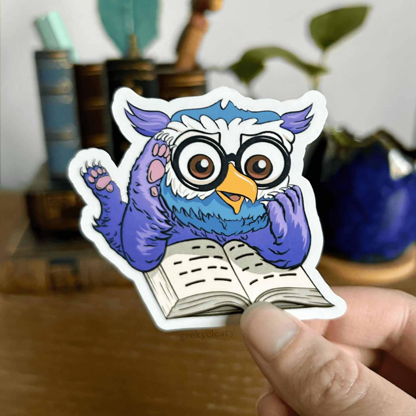 Fantasy owlbear shocked reading sticker for readers held by a hand above a wooden desk with books and a potted plant in the background.