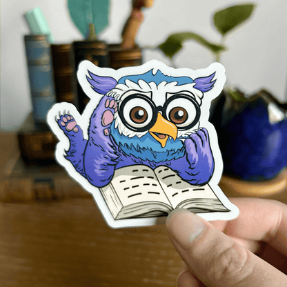 Fantasy owlbear shocked reading sticker for readers held by a hand above a wooden desk with books and a potted plant in the background.