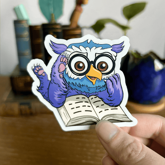 Fantasy owlbear shocked reading sticker for readers held by a hand above a wooden desk with books and a potted plant in the background.