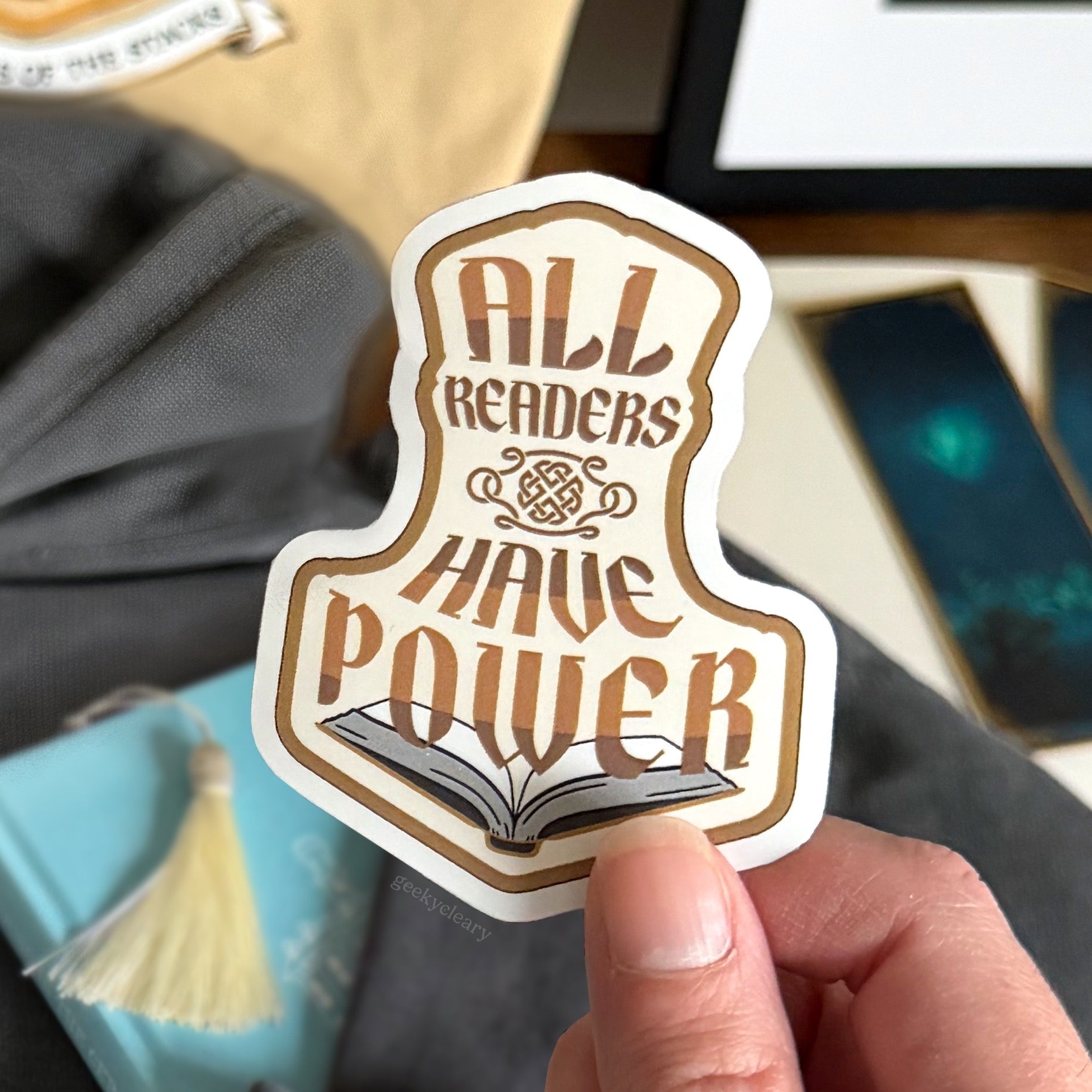Close up of All Readers Have Power vinyl sticker that readers can place anywhere they want their reader to show. Sticker held over bookish flat lay, books, and bookmarks.