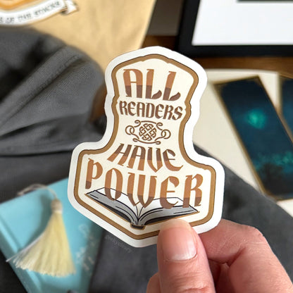 Close up of All Readers Have Power vinyl sticker that readers can place anywhere they want their reader to show. Sticker held over bookish flat lay, books, and bookmarks.