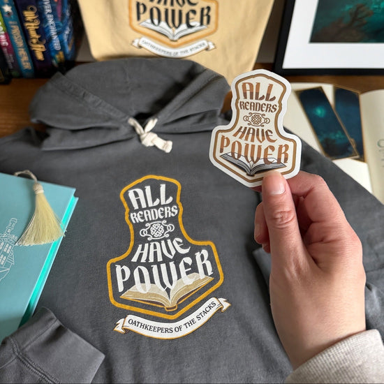 Hoodie with 'All Readers Have Power' design, tote bag, and matching sticker on desk with books.